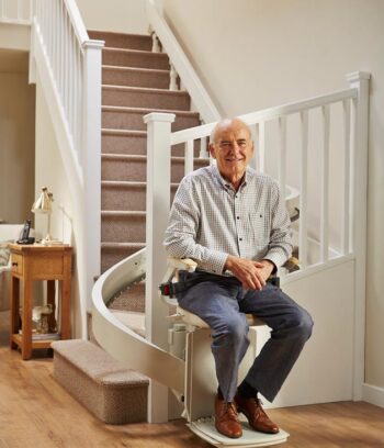 Elderly gentleman smiling, sat on chairlift at bottom of stairs.